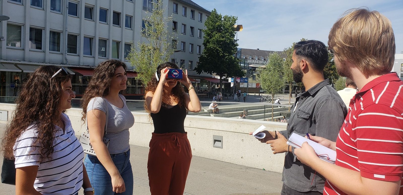 Field testing in a public square, a participant looks through a cardboard VR viewer while researchers take notes